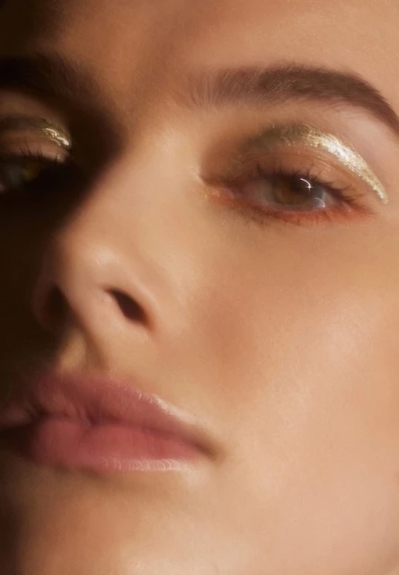 Close-up portrait of a woman with gold eyeshadow. Her face is partially visible, soft focus on skin texture and eye detail. 