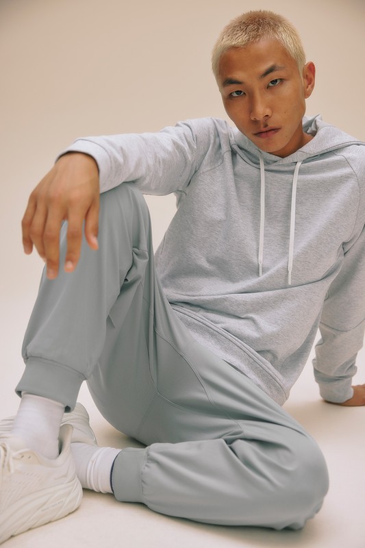 Man in gray hooded sweatshirt and athletic pants sitting on the floor. Portrait-style shot with a neutral backdrop and fashionable athletic wear.