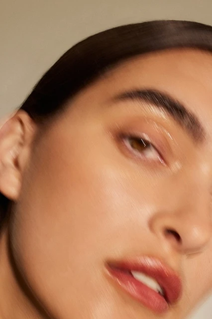 Close-up portrait of an adult female model with glossy makeup, dark hair, fair skin, and brown eyes, in a beauty or fashion context.