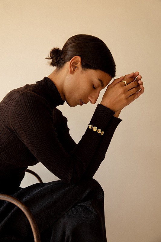 Profile portrait of a woman with dark hair in a bun wearing a black turtleneck and gold jewelry, seated in profile against a plain wall.
