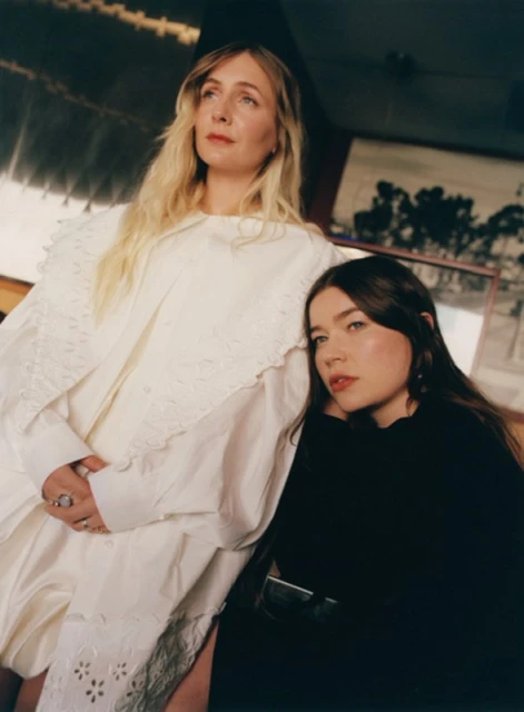 Two women pose indoors: one with long blonde hair in a white dress, the other with long brown hair in black. Soft lighting.