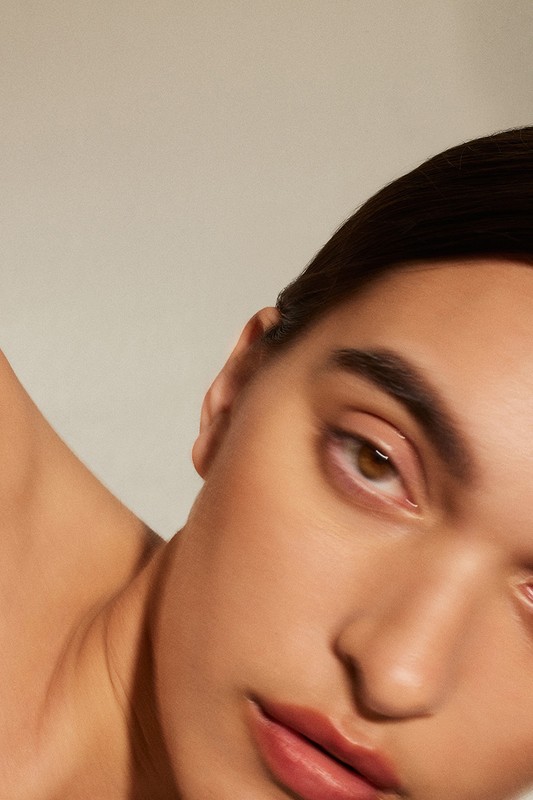 Close-up beauty portrait of a woman with natural makeup, dark hair, and brown eyes against a neutral background.