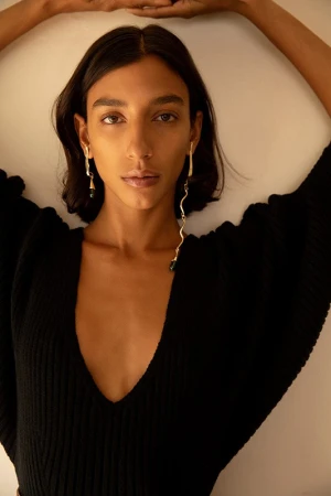 Portrait of a woman with long dark hair wearing a black V-neck ribbed top and gold drop earrings against a plain wall.