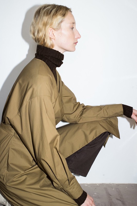 A blond adult female crouching indoors wearing an oversized khaki shirt and dark brown turtleneck in a fashion editorial shoot.