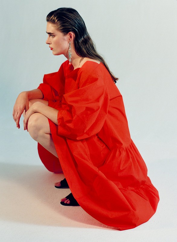 A solo fashion model poses in a bright red dress, squatting indoors under studio lighting, showcasing an editorial style and simple background.