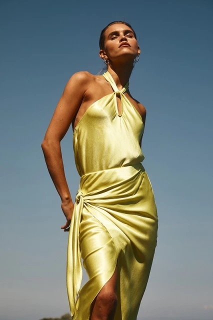 Woman in a yellow silk dress stands against a blue sky in a fashion shot, posing with her head tilted back and eyes closed.