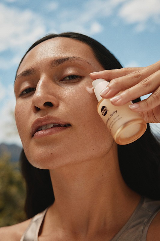 Close-up of a woman applying skincare from a pump bottle against a blue sky, trees and mountain background in natural light.