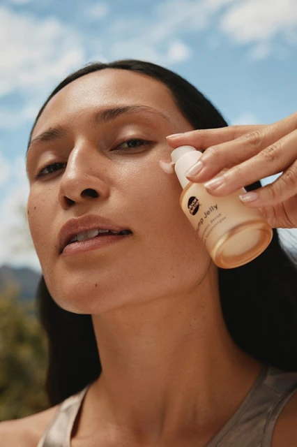 Close-up of a woman applying skincare from a pump bottle against a blue sky, trees and mountain background in natural light.
