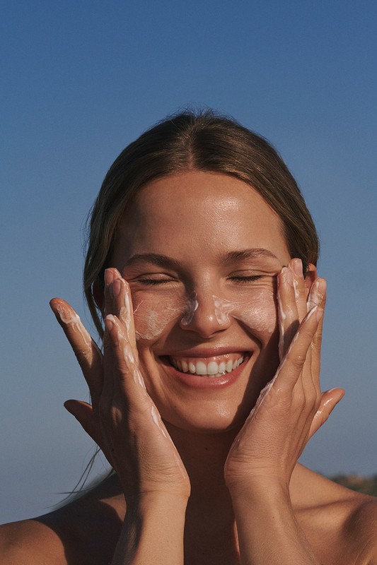 Smiling woman applying sunscreen to her face outdoors, eyes closed, bathed in sunlight against a clear blue sky in a close-up shot.