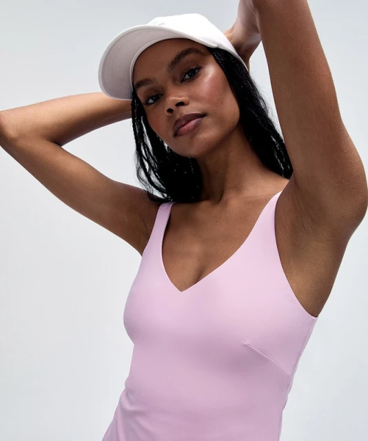 Close-up of a woman wearing a white baseball cap and a light purple tank top, with her arms raised behind her head in a studio setting.