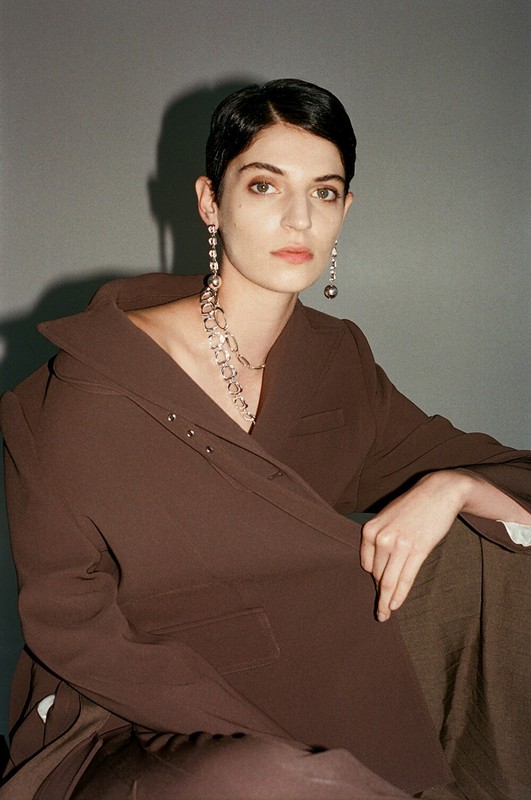 Elegant woman with short dark hair, wearing statement jewelry and a brown jacket and trousers, seated against a neutral background.
