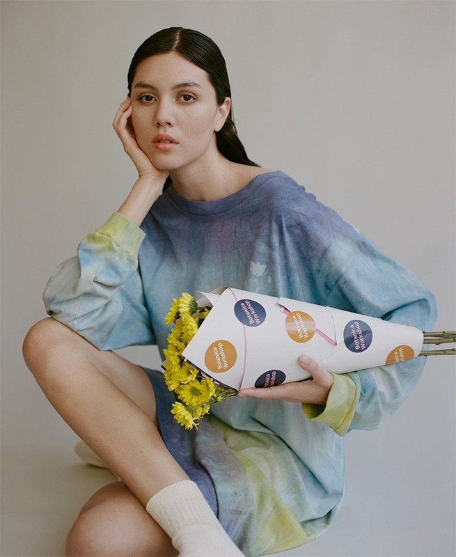 Portrait of a woman sitting with a bouquet of yellow flowers in a paper cone, wearing a blue tie-dye shirt and white socks against a grey backdrop.