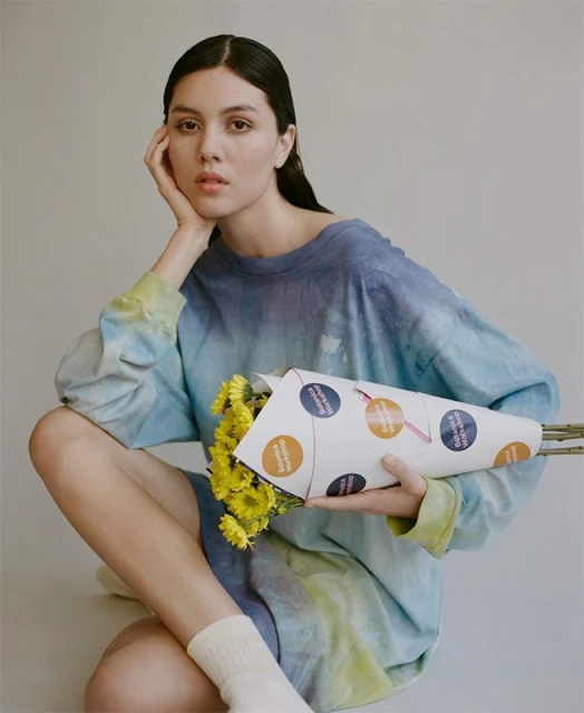 Portrait of a woman sitting with a bouquet of yellow flowers in a paper cone, wearing a blue tie-dye shirt and white socks against a grey backdrop.