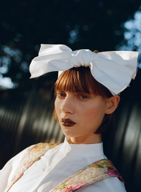 Portrait of a young woman with a large white bow in her hair, wearing a floral vest and white shirt, against a blurred, dark background.