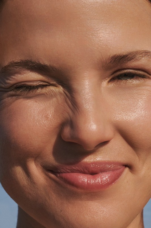 Close-up portrait of a smiling woman with glowing skin, one eye squinted, capturing a natural, joyful expression in bright, diffused light.