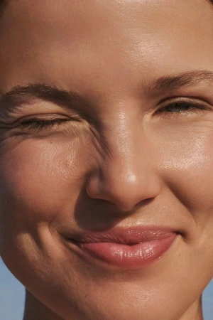 Close-up portrait of a smiling woman with glowing skin, one eye squinted, capturing a natural, joyful expression in bright, diffused light.
