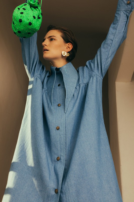 Adult woman in a light-blue, button-up shirt dress raising a green beaded bag with both hands in an indoor setting.