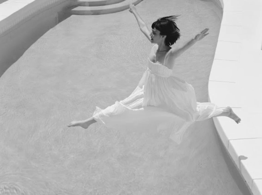 Black and white photograph of a woman in a white dress leaping over water in a pool with arms outstretched and legs extended.