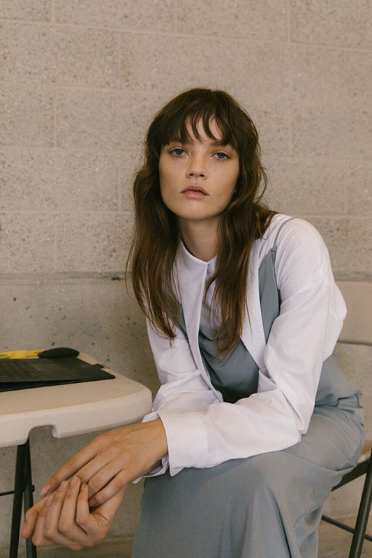 A woman with brown hair and bangs sits in an indoor setting, wearing a white shirt and grey dress, posing with her hands clasped.