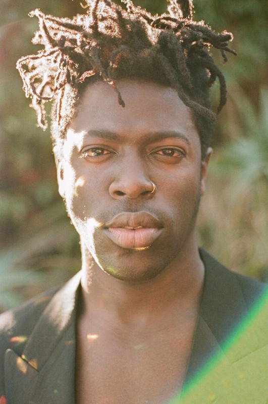 Close-up portrait of a Black man with dreadlocks wearing a black blazer outside with natural light creating a lens flare effect.