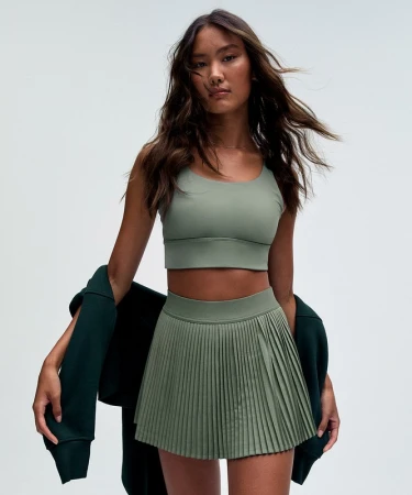Portrait of an East Asian woman wearing a sage green athletic crop top and pleated tennis skirt, against a seamless white background.