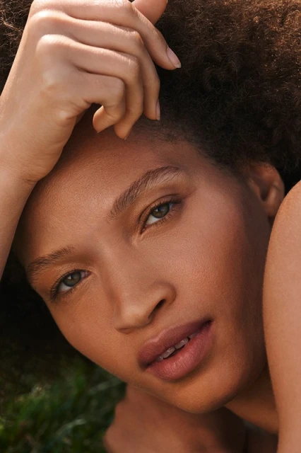 Close-up portrait of a woman with dark curly hair and tan skin, posing outdoors with green foliage in the background on a sunny day.