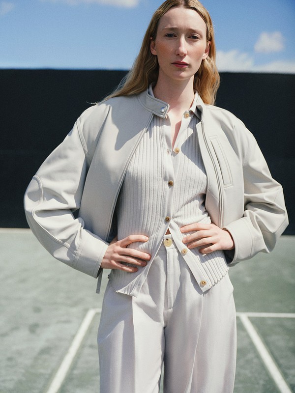 A woman in a light grey outfit poses on a tennis court, wearing a jacket, ribbed shirt, and trousers with her hands on her hips.