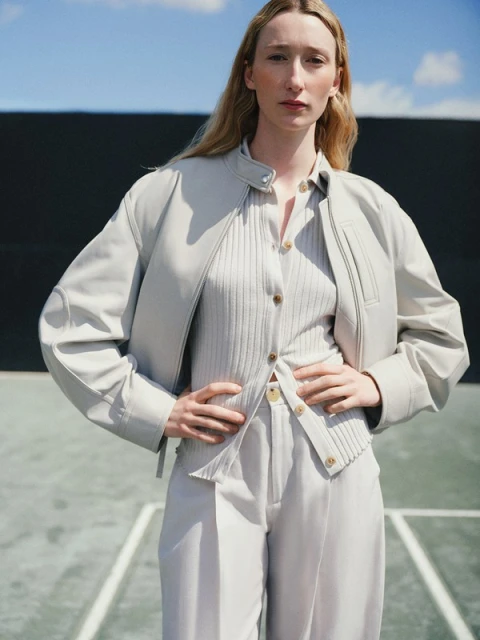 A woman in a light grey outfit poses on a tennis court, wearing a jacket, ribbed shirt, and trousers with her hands on her hips.