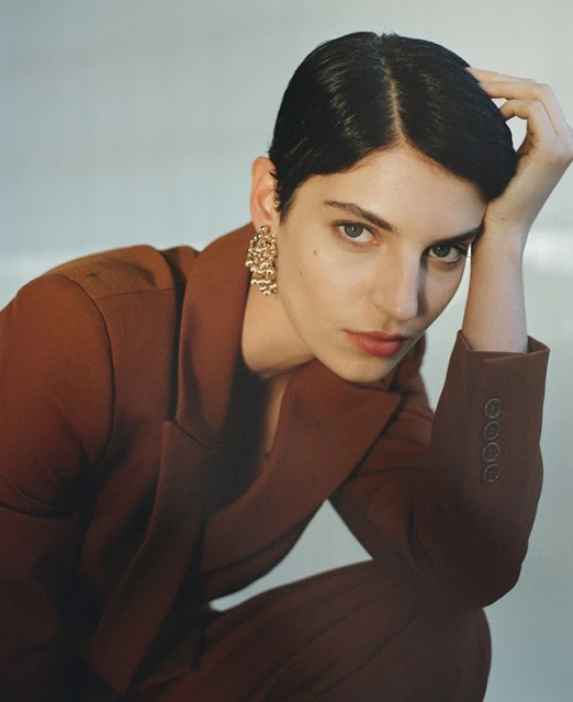Portrait of a fashionable adult woman in a rust-colored suit with short, dark hair and gold earrings against a neutral backdrop.