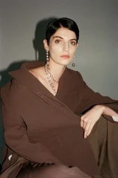 Model with dark short hair wearing a brown blazer and large silver earrings against a plain grey background