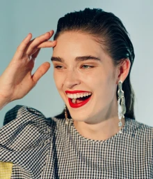 Close-up portrait of a smiling woman with slicked-back hair wearing a gingham top and bold red lipstick against a light background.