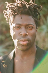 Close-up portrait of a black man with locs, nose ring, and dark jacket against a softly blurred natural background.
