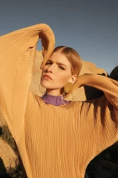 A woman in a pleated gold top and large hoop earrings posing outdoors against large desert rock formations and blue sky.