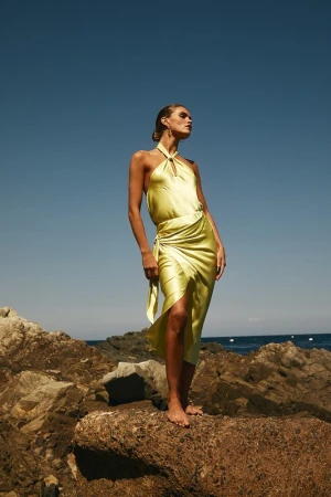 Model in a yellow satin halter dress standing on rocky coastal terrain against a clear blue sky