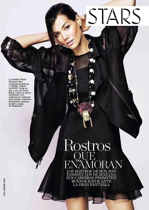 Editorial fashion shot of a woman wearing a black dress and a large pendant necklace, posing with her hands up near her head.