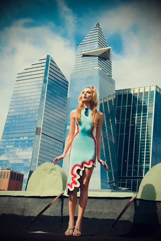 Young woman poses in a unique dress against a modern cityscape backdrop with blue sky and clouds, creating a striking fashion image.