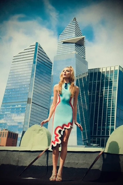 Young woman poses in a unique dress against a modern cityscape backdrop with blue sky and clouds, creating a striking fashion image.