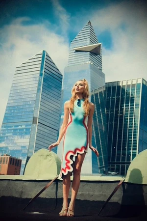 Young woman poses in a unique dress against a modern cityscape backdrop with blue sky and clouds, creating a striking fashion image.