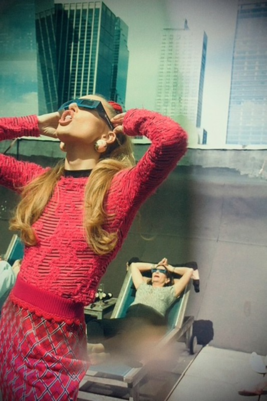 Two women wearing sunglasses pose on a rooftop in front of city buildings. One stands in a red outfit, the other reclines in a chair.