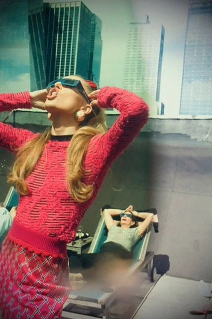 Two women wearing sunglasses pose on a rooftop in front of city buildings. One stands in a red outfit, the other reclines in a chair.