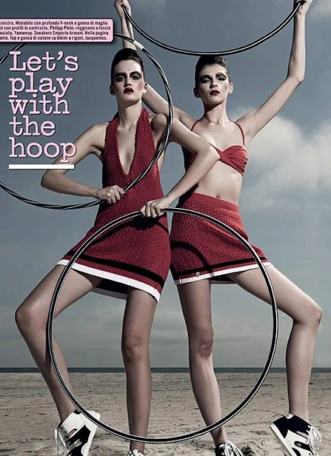Two fashion models posing on a beach holding hula hoops, wearing red knit mini skirts and tops, sporty sneakers, dramatic eye makeup.