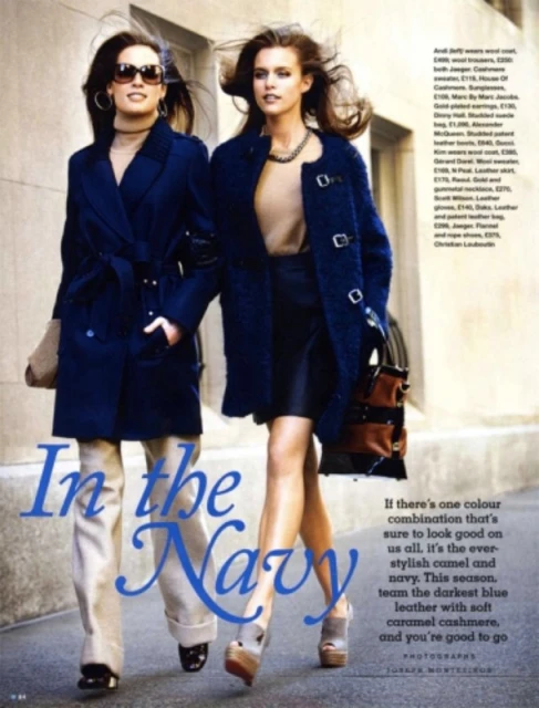 Two women model navy blue coats in a fashion editorial. The blonde woman carries a brown handbag and wears platform heels.