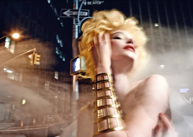 Adult female with curly blonde hair poses with gold cuff bracelet in blurry urban nighttime street scene with street sign and traffic light.