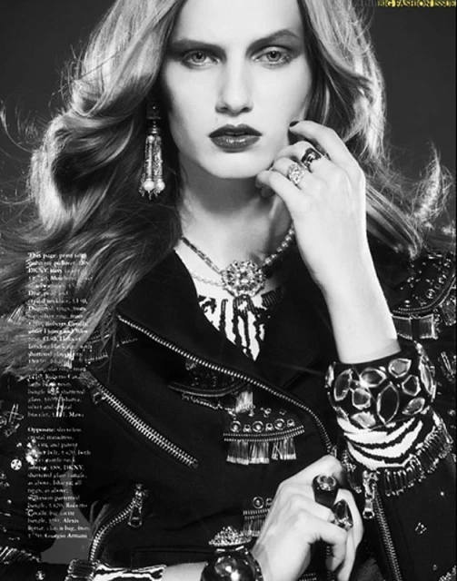 Black and white fashion portrait of a model wearing embellished leather jacket, jewelry, rings, and dark lipstick against a gray background.