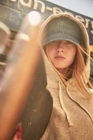 Close-up portrait of a woman wearing a grey hoodie and denim baseball cap holding a skateboard in Los Angeles.