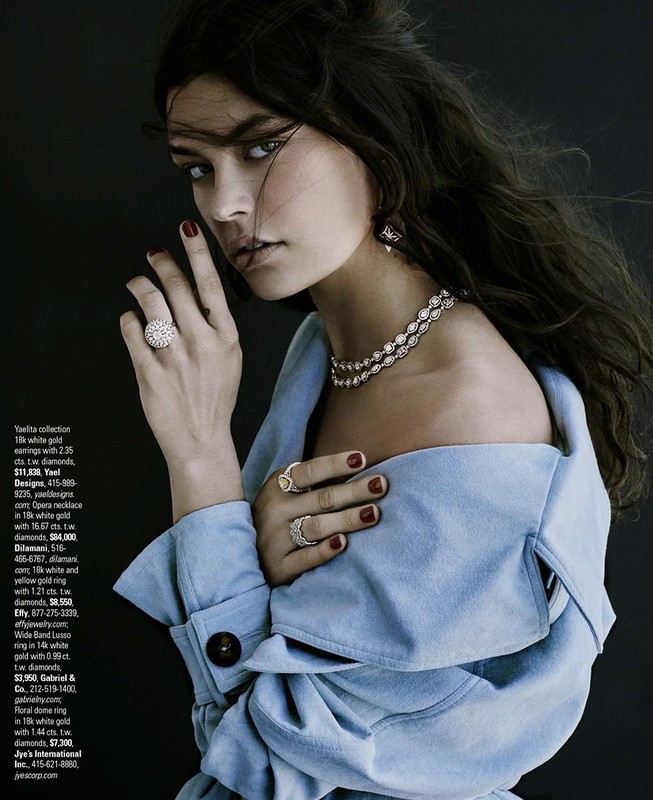 Glamorous portrait of a woman wearing a blue jacket and diamond jewelry including earrings, a ring, and a necklace against a black background.