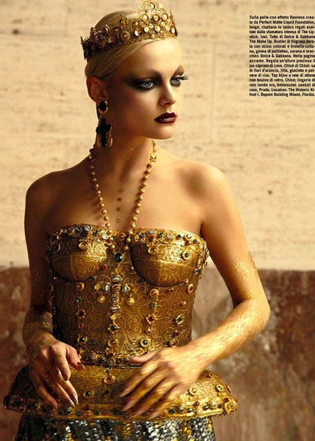 Fashion portrait of a blond woman wearing an ornate gold crown and jeweled bodice against a neutral background in soft studio light.