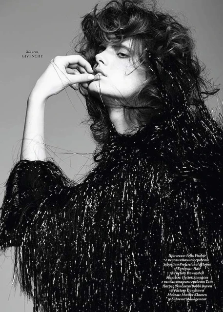 Monochrome close-up of a young adult woman with curly hair, wearing a black fringed top, with her fingers near her mouth.