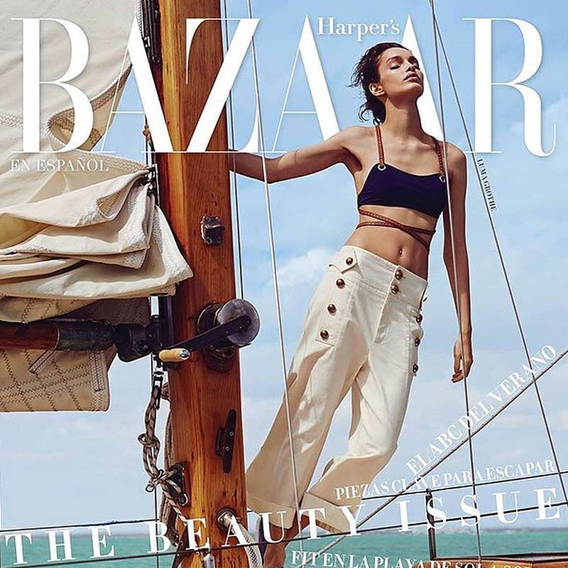 Magazine cover of Harper's Bazaar featuring a model in a navy bikini top and white pants posing on a sailboat with an ocean background.