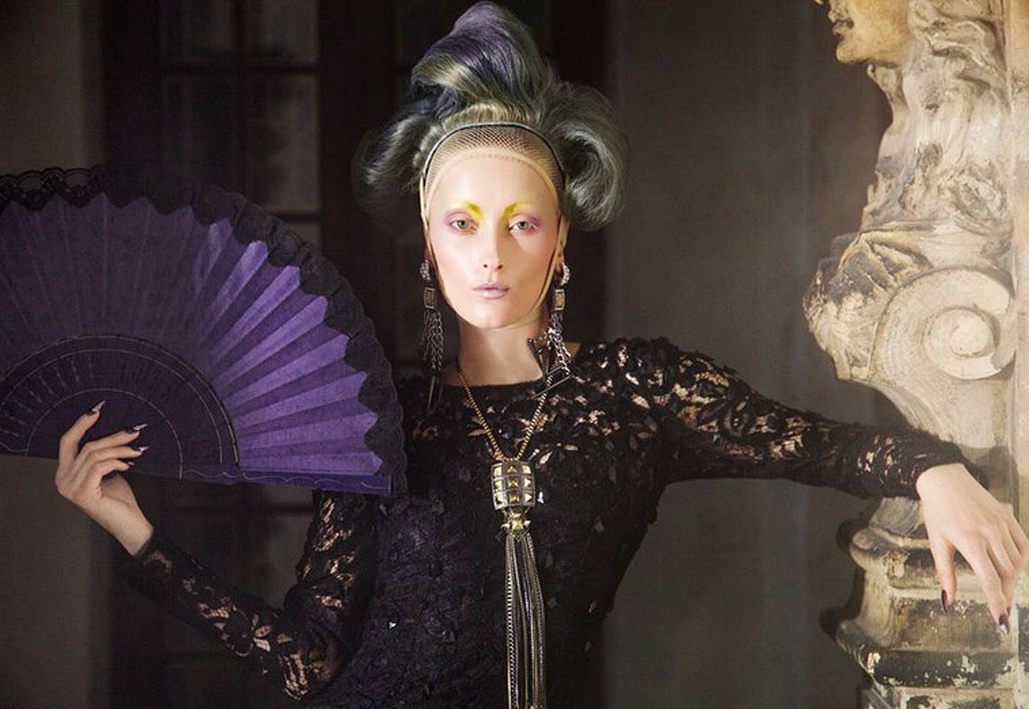 Elegant woman in black lace dress and elaborate makeup poses with a purple hand fan in an indoor setting with a classical architectural backdrop.